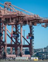 Three containers hang from cranes at Terminal 46 in Seattle. The middle container has a “Welcome to the Pacific Northwest” message to FIFA World Cup visitors, while the outside containers have a United State and Australian flag.