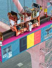 A shipping container with a Belgium flag banner hangs from a crane at Terminal 46 in Seattle.
